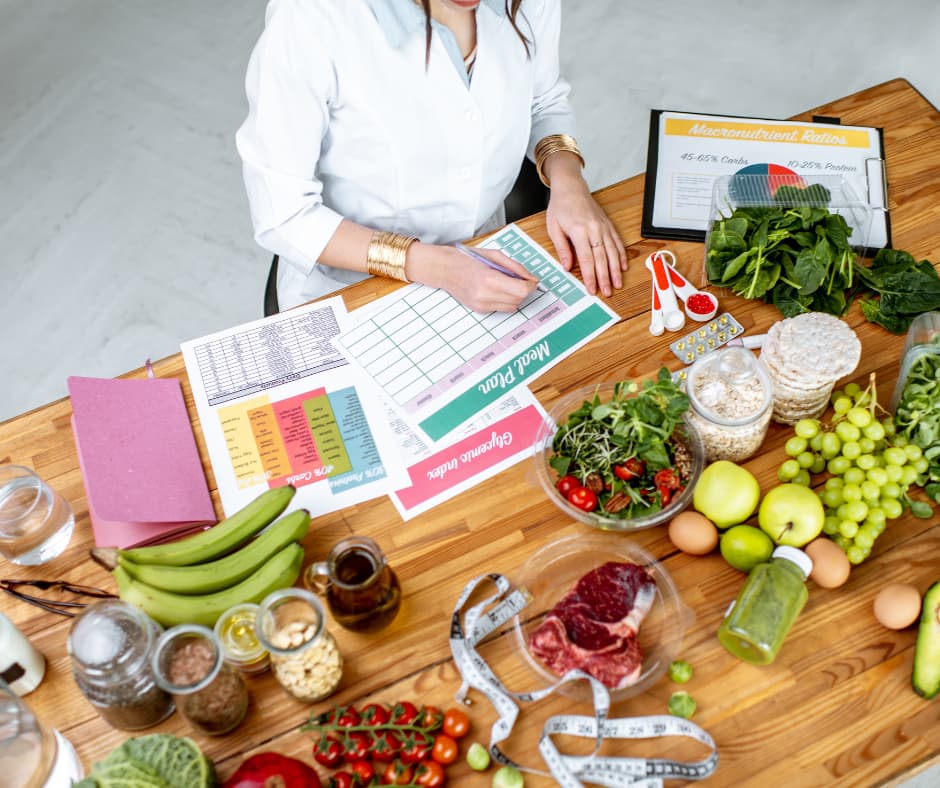 Nutritionist writing a meal plan at a table covered with fresh produce, supplements, and a macronutrient chart